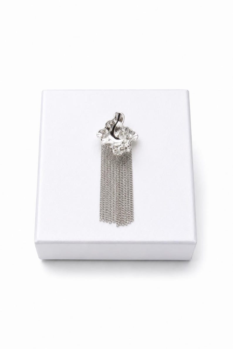 May include: A silver brooch with a textured, organic-shaped top and a cascade of dangling chains. The brooch is displayed on a white surface, creating a contrast that highlights its metallic sheen. The design is modern and elegant.