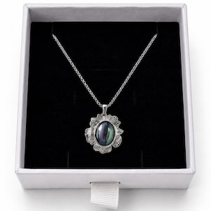 Pode incluir: Um colar de tom prateado com um pendente de pedra escura iridescente de forma oval, inserido numa moldura de metal texturizada e recortada. O pendente pende de uma delicada corrente prateada, apresentado numa caixa branca quadrada.