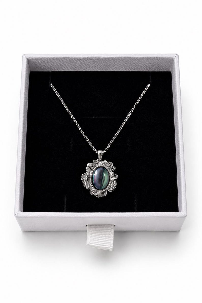 Pode incluir: Um colar de tom prateado com um pendente de pedra escura iridescente de forma oval, inserido numa moldura de metal texturizada e recortada. O pendente pende de uma delicada corrente prateada, apresentado numa caixa branca quadrada.