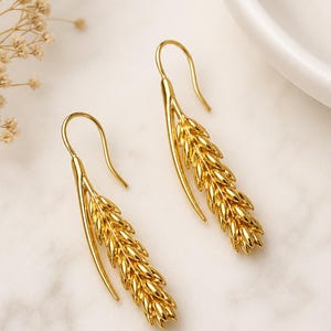 May include: A pair of gold-colored earrings in the shape of wheat stalks. The earrings have a hook design for pierced ears. The wheat stalk design is detailed and textured, with a shiny finish. The earrings are displayed on a white marble surface.