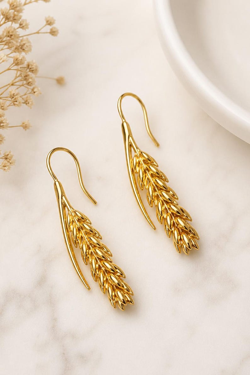 May include: A pair of gold-colored earrings in the shape of wheat stalks. The earrings have a hook design for pierced ears. The wheat stalk design is detailed and textured, with a shiny finish. The earrings are displayed on a white marble surface.