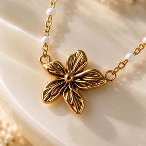May include: A gold-toned necklace featuring a central five-petaled flower pendant with textured detailing. The chain is composed of thin gold-colored metal links interspersed with small, round, white beads that create a delicate and refined appearance.