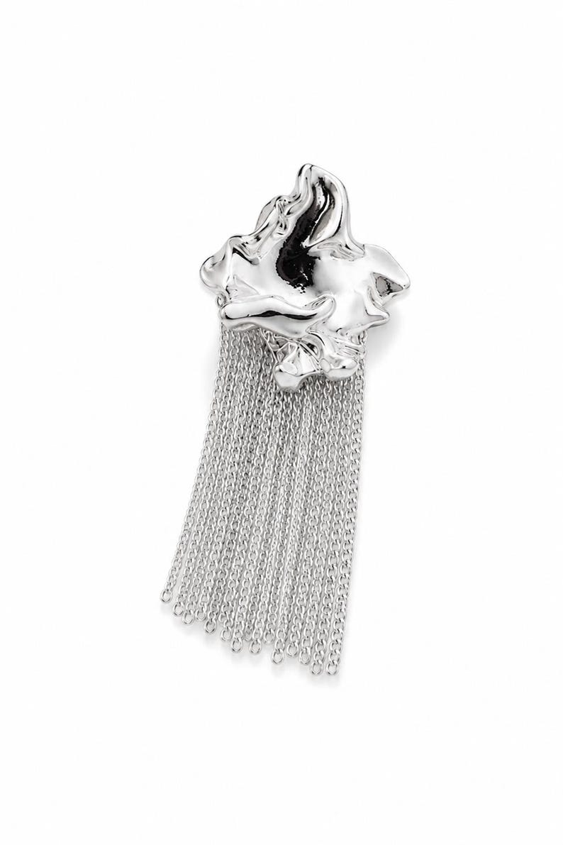 May include: A silver brooch with a fluid, abstract design and a fringe of fine chains. The polished surface reflects light, offering a contemporary and stylish accessory. The brooch is a statement piece.