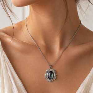 Pode incluir: Um colar de tom prateado com um pendente que apresenta uma pedra central oval de cor escura iridescente. A pedra &eacute; rodeada por uma moldura de prata texturizada em forma de p&eacute;talas. O pendente est&aacute; suspenso numa delicada corrente prateada.