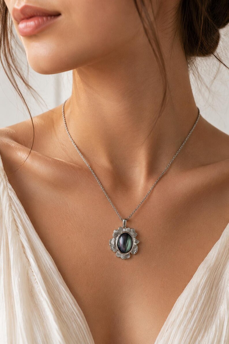 Pode incluir: Um colar de tom prateado com um pendente que apresenta uma pedra central oval de cor escura iridescente. A pedra &eacute; rodeada por uma moldura de prata texturizada em forma de p&eacute;talas. O pendente est&aacute; suspenso numa delicada corrente prateada.