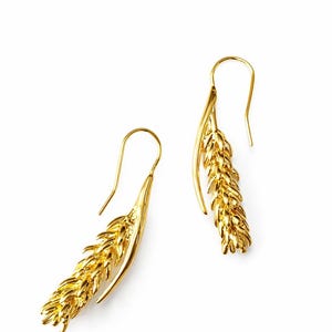 May include: A pair of gold-colored earrings in the shape of wheat stalks. The earrings have a hook design for pierced ears. The wheat stalks are detailed with textured surfaces, and the earrings are set against a white background.