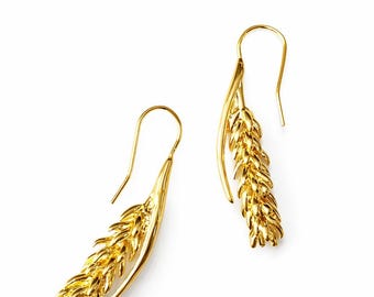 May include: A pair of gold-colored earrings in the shape of wheat stalks. The earrings have a hook design for pierced ears. The wheat stalks are detailed with textured surfaces, and the earrings are set against a white background.