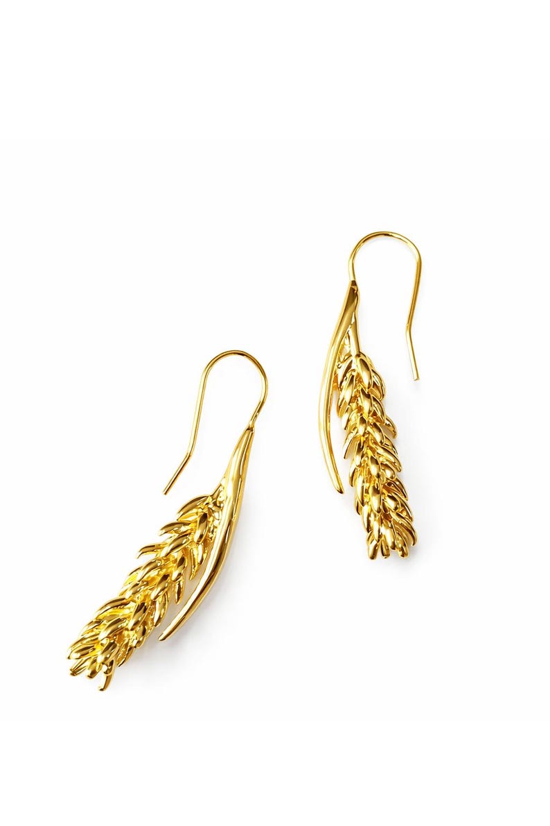 May include: A pair of gold-colored earrings in the shape of wheat stalks. The earrings have a hook design for pierced ears. The wheat stalks are detailed with textured surfaces, and the earrings are set against a white background.
