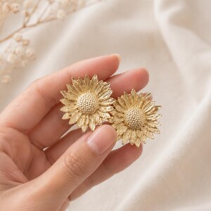 May include: A pair of gold-coloured sunflower earrings. Each earring has a detailed, textured design mimicking the petals and centre of a sunflower. The earrings are held in a hand against a soft, neutral background, showcasing the jewellery's intricate details.