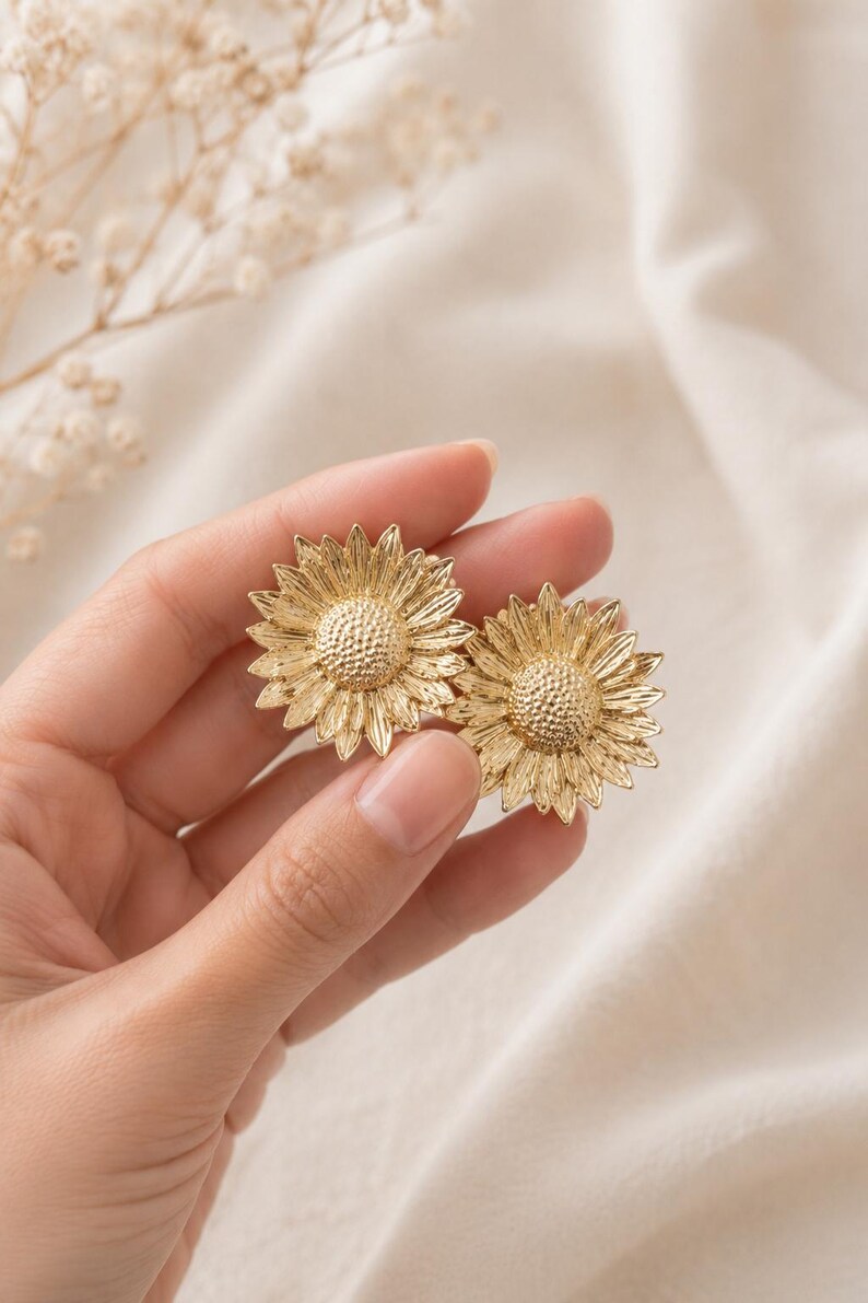 May include: A pair of gold-coloured sunflower earrings. Each earring has a detailed, textured design mimicking the petals and centre of a sunflower. The earrings are held in a hand against a soft, neutral background, showcasing the jewellery's intricate details.
