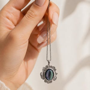 Pode incluir: Um colar de tom prateado com um pendente de pedra escura iridescente de formato oval, inserido numa moldura de metal texturizada inspirada num sol. O pendente pende de uma corrente fina e delicada de cor prateada com acabamento polido.