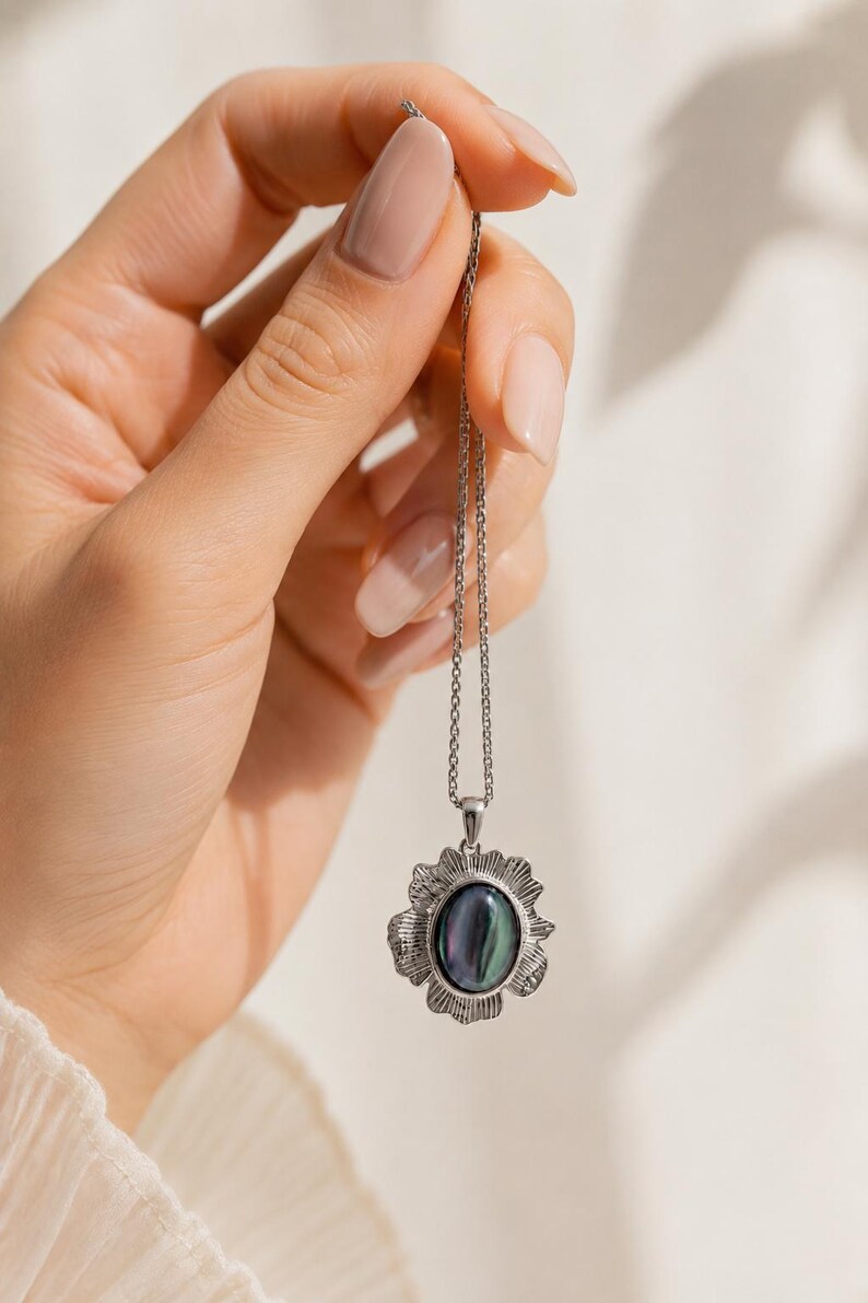 Pode incluir: Um colar de tom prateado com um pendente de pedra escura iridescente de formato oval, inserido numa moldura de metal texturizada inspirada num sol. O pendente pende de uma corrente fina e delicada de cor prateada com acabamento polido.