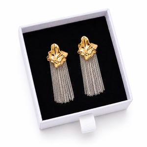 May include: A pair of gold-colored earrings with a textured, organic shape and dangling silver chain fringe. The earrings are displayed in a white box with a black interior.