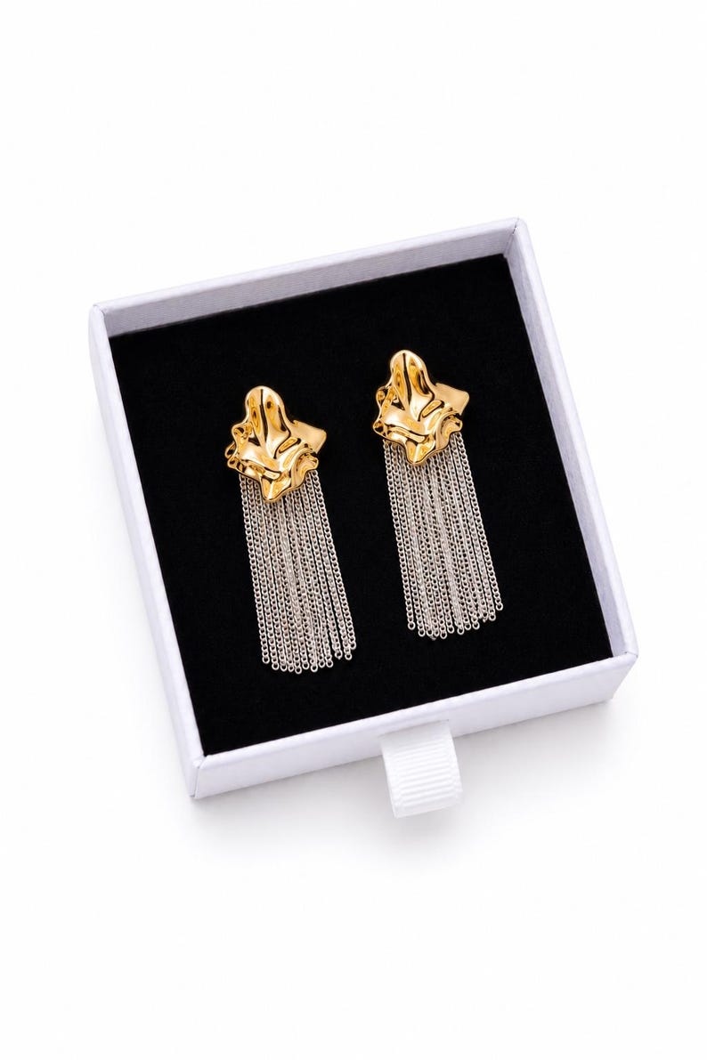 May include: A pair of gold-colored earrings with a textured, organic shape and dangling silver chain fringe. The earrings are displayed in a white box with a black interior.