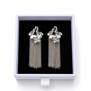 May include: Silver earrings with a floral design and chain fringe, presented in a white box with a black interior. The earrings have a textured, organic shape. The chains hang vertically, creating a flowing effect. The box is on a white background.