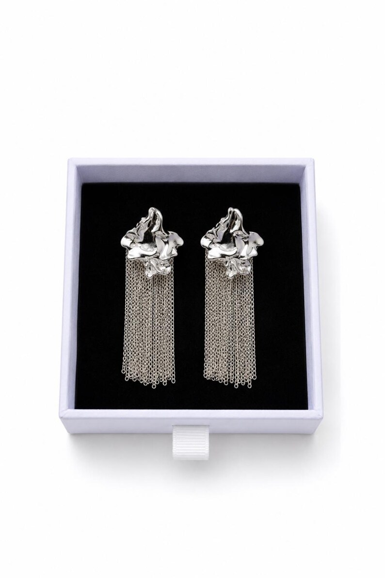 May include: Silver earrings with a floral design and chain fringe, presented in a white box with a black interior. The earrings have a textured, organic shape. The chains hang vertically, creating a flowing effect. The box is on a white background.