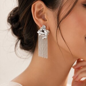 May include: Silver earrings with an abstract, textured design. Each earring has a sculpted element at the ear, with multiple strands of fine chain hanging down. These earrings are a contemporary accessory, perfect for adding a touch of elegance.