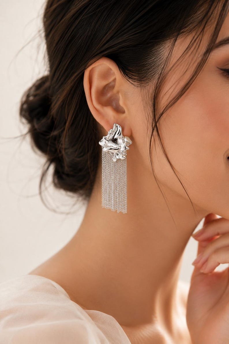 May include: Silver earrings with an abstract, textured design. Each earring has a sculpted element at the ear, with multiple strands of fine chain hanging down. These earrings are a contemporary accessory, perfect for adding a touch of elegance.
