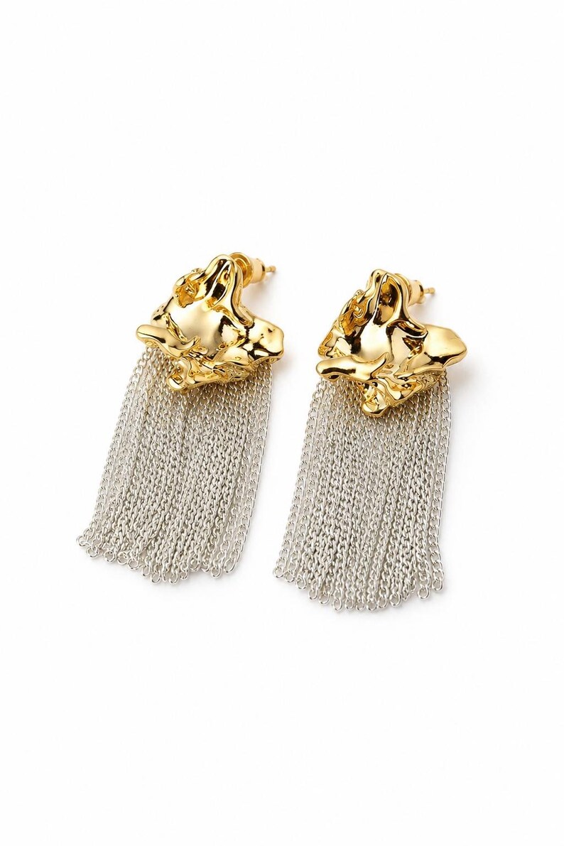 May include: A pair of gold-colored earrings with a textured, organic design. Each earring features a cascade of silver-colored chain fringe, creating a striking contrast. The earrings are presented against a clean white background.