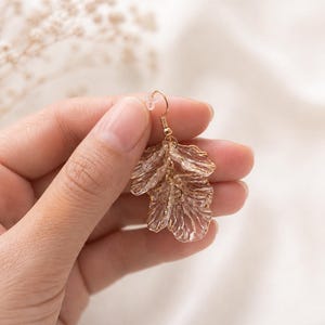 May include: A gold-colored earring with a floral design. The earring is held up by a gold-colored hook. The floral design is made of a translucent material with a textured surface. The background is a soft, neutral color.