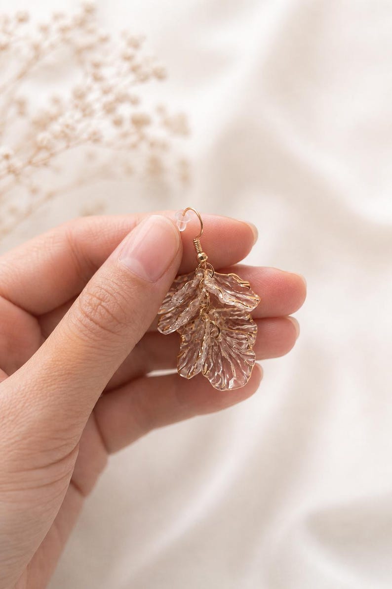 May include: A gold-colored earring with a floral design. The earring is held up by a gold-colored hook. The floral design is made of a translucent material with a textured surface. The background is a soft, neutral color.