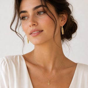 May include: A woman wearing gold jewelry, including a necklace with a small pendant and matching earrings. The earrings have a leaf-like design. The woman is wearing a white top.