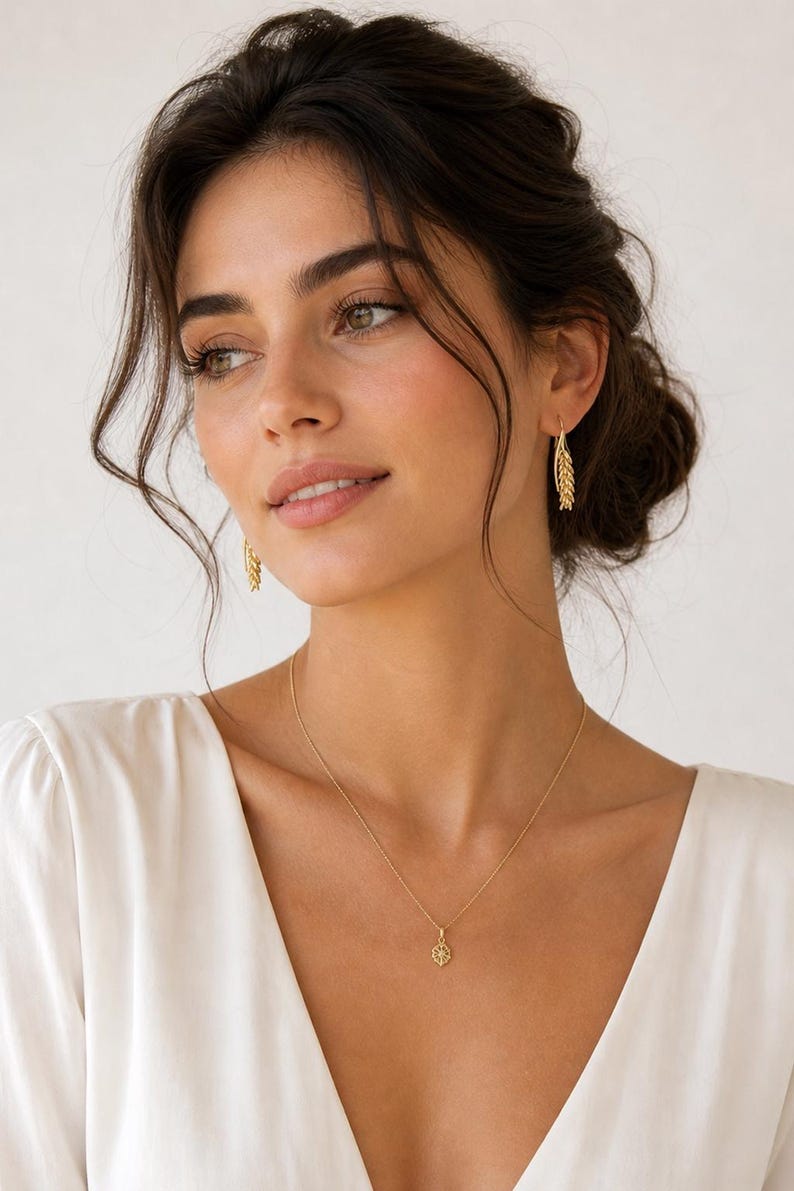 May include: A woman wearing gold jewelry, including a necklace with a small pendant and matching earrings. The earrings have a leaf-like design. The woman is wearing a white top.