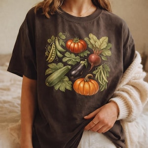 May include: A dark charcoal gray short-sleeve t-shirt featuring a central graphic illustration of various garden vegetables including a red tomato, a purple eggplant, an orange pumpkin, green peas, a zucchini, and a beet with green leaves.