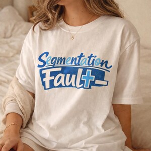 Può includere: Una t-shirt bianca a maniche corte con la scritta "Segmentation Fault" stampata in blu e azzurro. La parola "Segmentation" è in corsivo, mentre "Fault" è in un carattere grassetto su uno sfondo rettangolare di colore blu.