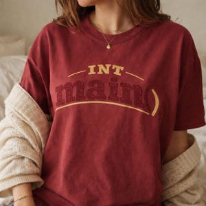 May include: A maroon short-sleeved t-shirt featuring the text INT main() in a gold and dark red glitter font. The shirt has a crew neckline and is layered with a cream-colored knitted cardigan draped over the shoulders, showcasing a casual style.
