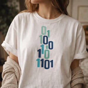 May include: A white short-sleeved t-shirt featuring a vertical graphic print of binary code digits in shades of light blue, teal, and dark blue. The numbers 0 and 1 are arranged in a stacked, staggered pattern across the center of the chest area.