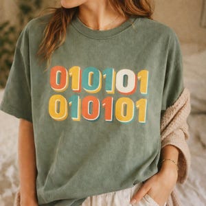 May include: A sage green short-sleeve t-shirt featuring the binary code sequence 010101 printed in two rows. The numbers are rendered in a retro-style font with layered colors of orange, teal, yellow, and white, creating a bold graphic design.