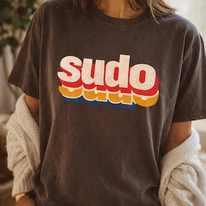 Op de afbeelding: Een antracietgrijs T-shirt met korte mouwen en het woord "sudo" in een vet, wit, verweerd lettertype. Onder de hoofdtekst verschijnen twee extra gelaagde, versprongen schaduwen in levendig rood, geel en blauw, wat een gestapeld effect geeft.