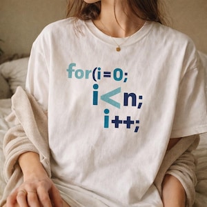 Könnte beinhalten: Ein weißes kurzärmeliges T-Shirt mit einem blauen und hellblauen Grafikdruck eines Programmier-For-Schleifen-Code-Schnipsels, der for(i=0; i<n; i++); lautet. Das T-Shirt hat einen Rundhalsausschnitt und wird mit einer beigen, strukturierten Strickjacke getragen.
