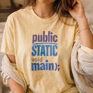 May include: A pale yellow short-sleeved t-shirt featuring the dark blue and light blue text "public STATIC void main);" printed on the front. The shirt has a crew neckline and a relaxed fit, showcasing a programming-themed graphic design.