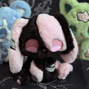 May include: A plush bunny toy with black and pink fur, embroidered facial features, and bandages. The bunny has long ears and is sitting. Other plush toys in blue and green are in the background.
