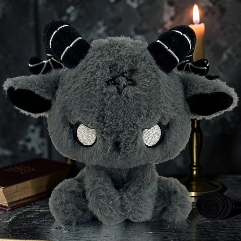 May include: A plush, gray goat-like creature with black horns and ears, and white eyes. The stuffed animal has a pentagram design on its forehead and is sitting with its paws clasped. A lit candle and a book are in the background.