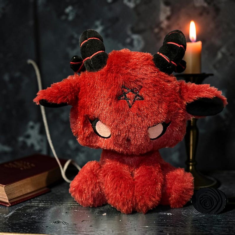 May include: A plush red goat-like creature with black horns and a pentagram on its chest. The stuffed animal has pink accents around the eyes and a fluffy texture. A lit candle and a book are in the background.