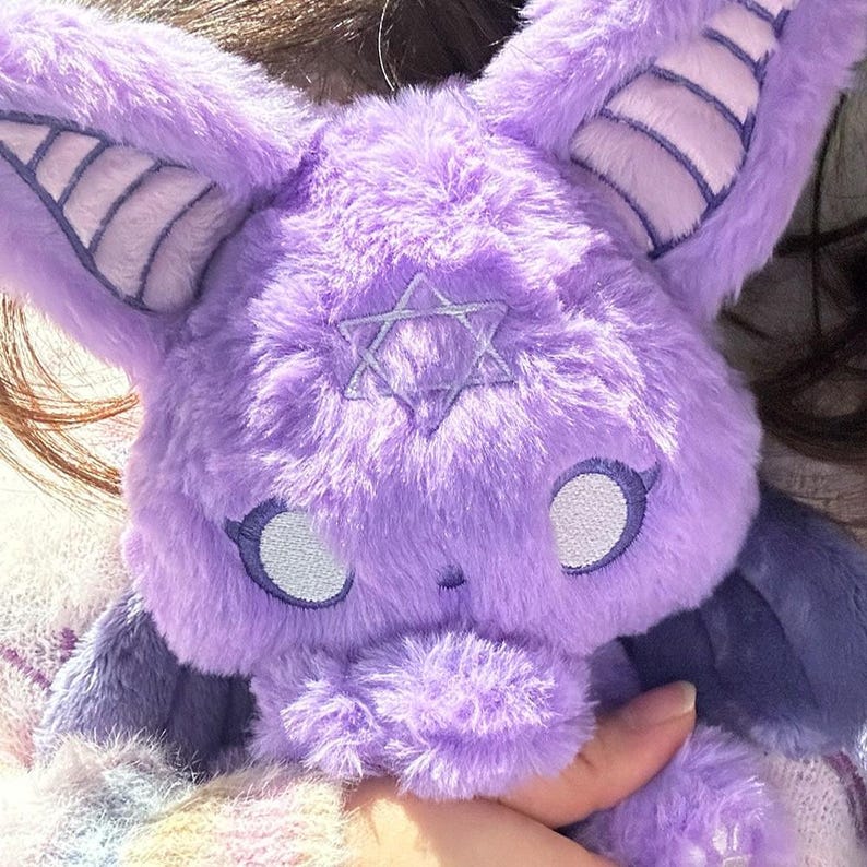 May include: A plush, lavender-colored creature with large, pointed ears and a star emblem on its chest. The creature has light blue accents on its ears and around its eyes. The plush toy is soft and fluffy.