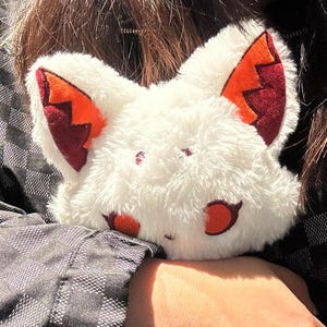 May include: A white plush fox-shaped accessory with orange and burgundy accents on the ears and eyes. The fluffy material creates a soft texture. The accessory is designed to be worn or held.