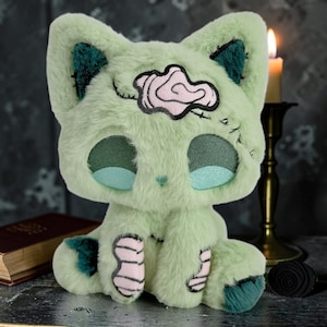 Può includere: Un gatto di peluche verde chiaro con dettagli verde scuro sulle orecchie e sulle zampe. Il gatto ha un disegno rosa e nero sulla fronte e dettagli cuciti. Gli occhi sono verde scuro con centri azzurri. Il gatto è seduto.