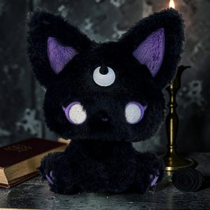 May include: A black plush cat toy with purple inner ears, eyebrows, and paw pads. The cat features white eyes and a third eye on its forehead. The toy is sitting upright and has a fluffy texture.