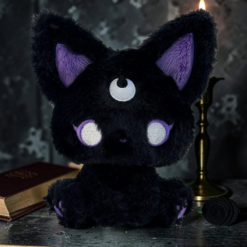 May include: A black plush cat with purple inner ears, eyebrows, and paw pads. The cat has white eyes and a third eye on its forehead. The plush toy is sitting upright, with a fluffy texture.
