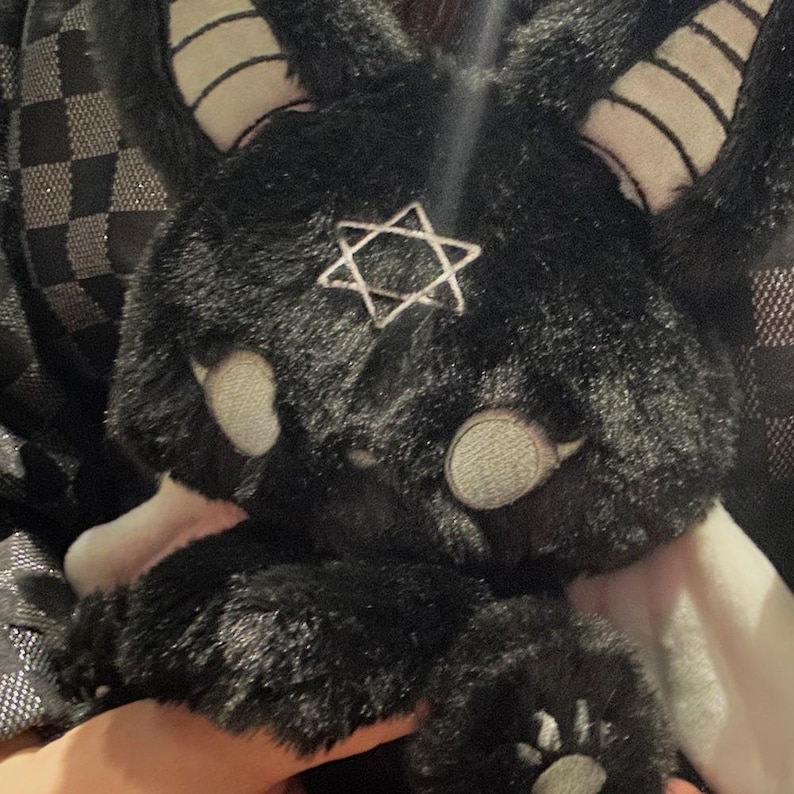 May include: A black plush bunny with white accents, including horns, eyes, and paws. A silver star is attached to the bunny's chest. The bunny is a unique, handmade item.