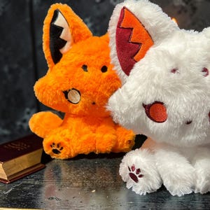 May include: Two plush fox toys, one orange and one white, sit side-by-side. The orange fox has black accents and a white inner ear. The white fox has red accents. Both have paw print details on their feet. A small book is visible in the background.