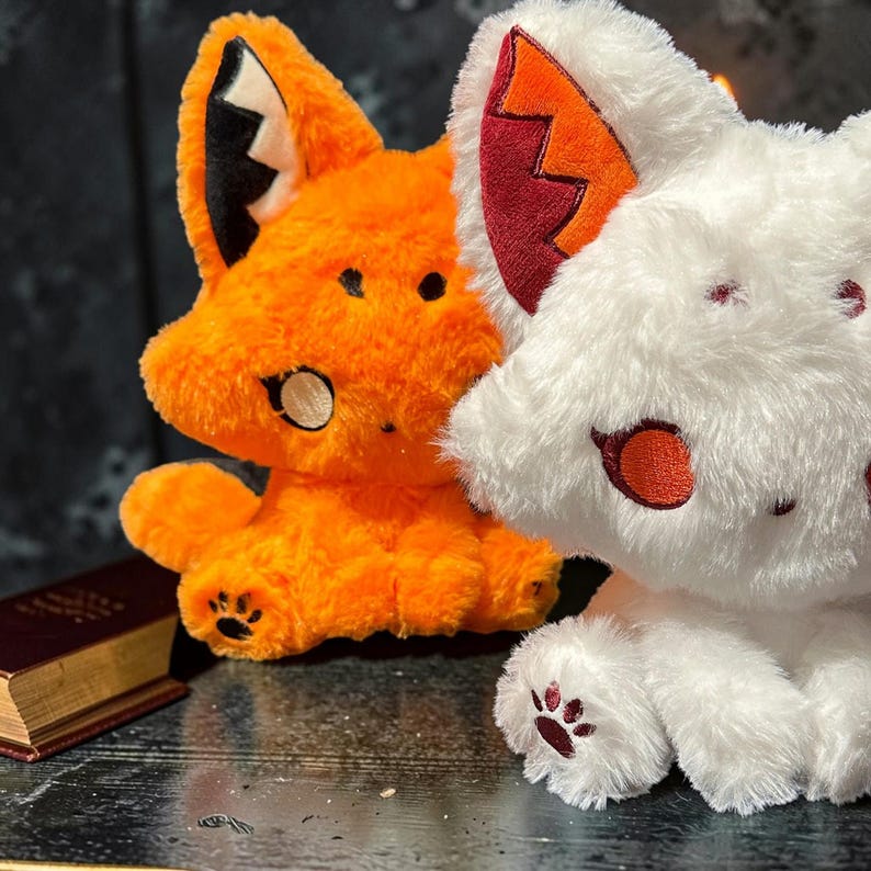 May include: Two plush fox toys, one orange and one white, sit side-by-side. The orange fox has black accents and a white inner ear. The white fox has red accents. Both have paw print details on their feet. A small book is visible in the background.