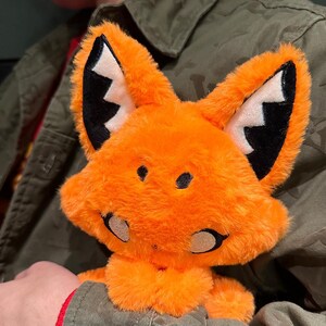 May include: A plush orange fox toy with black and white accents on the ears and face. The fox has a friendly expression with black eyes and a stitched nose. The toy is made of soft, fuzzy material.