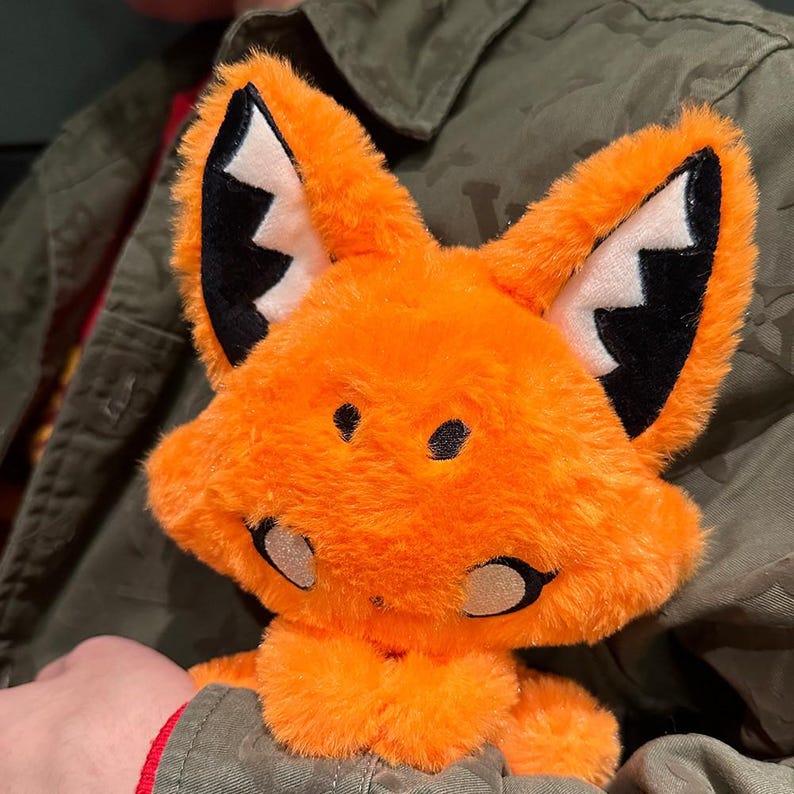 May include: A plush orange fox toy with black and white accents on the ears and face. The fox has a friendly expression with black eyes and a stitched nose. The toy is made of soft, fuzzy material.