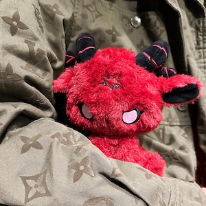 May include: A plush red devil doll with black horns and a star on its forehead. The doll has pink accents around the eyes and mouth. The doll is held against a green jacket with a repeating floral pattern.