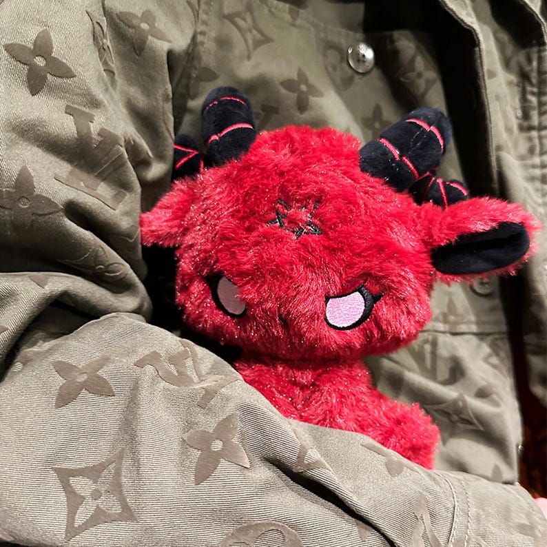 May include: A plush red devil doll with black horns and a star on its forehead. The doll has pink accents around the eyes and mouth. The doll is held against a green jacket with a repeating floral pattern.